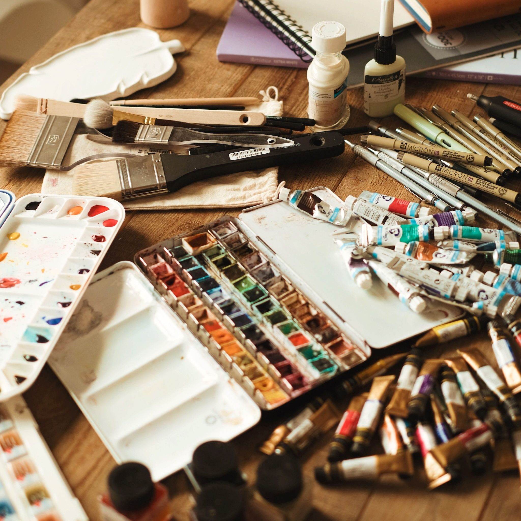 Art supplies spread across a studio table — watercolor palettes, paint tubes, brushes, and pens