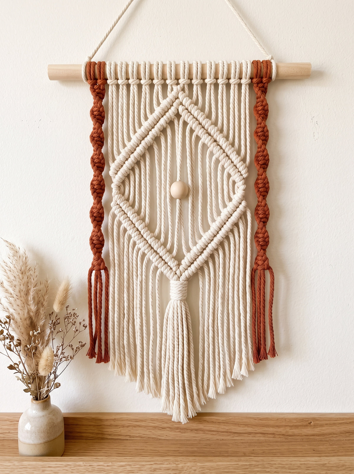 Cream and rust macramé wall hanging with a diamond pattern and wood bead on a wooden dowel