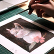 Gouache portrait of a white cat in progress with the artist's brush in hand